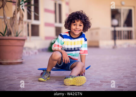 Kleiner Junge mit lockigem Haar mit Skateboard outdoor. Kinder und Unterhaltung Konzept Stockfoto