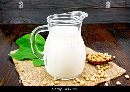 Sojamilch in einem Krug, frische grüne Blätter, Sojabohnen in einem Löffel und sackleinen auf dem Hintergrund einer dunklen Holzplatte Stockfoto