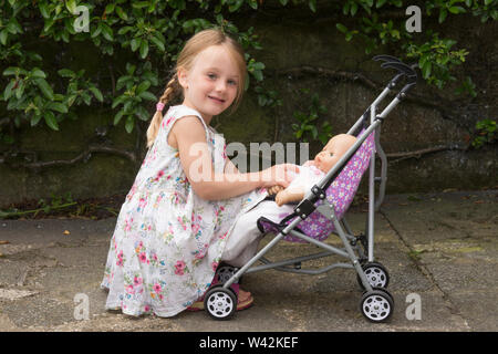 Drei Jahre alten Kind, junge Mädchen in der hübschen Kleid, blonde Haare in Pigtails spielen mit Puppe in Spielzeug Kinderwagen, draußen im Garten, Großbritannien Stockfoto