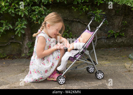 Drei Jahre alten Kind, junge Mädchen in der hübschen Kleid, blonde Haare in Pigtails spielen mit Puppe in Spielzeug Kinderwagen, draußen im Garten, Großbritannien Stockfoto