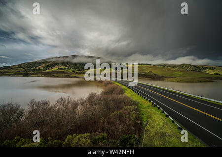 Leere Straße zwischen zwei Stauseen mit grünen Hügeln und Gewitterwolken Stockfoto