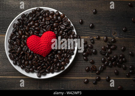 Rotes Herz auf eine Menge von gerösteten Kaffeebohnen Hintergrund auf weiße Platte auf hölzernen Tisch. Starken schwarzen Kaffee Espresso, Körner, Hintergrund, Textur Stockfoto