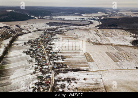 Blick von oben auf die leeren verschneite Felder, Häuser entlang der Straße und bewaldeten Hügeln auf und blauer Himmel. Antenne drone Fotografie, Winterlandschaft. Stockfoto