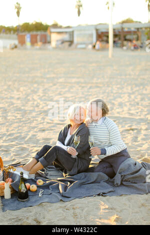 Zwei glückliche Rentner sitzen auf Plaid auf Sand Strand mit Champagner und Obst. Konzept des älteren Paares auf Picknick. Stockfoto