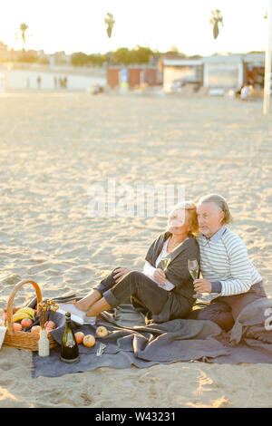 Zwei Rentner sitzen auf Plaid auf Sand Strand mit Champagner und Obst. Konzept des älteren Paares auf Picknick. Stockfoto