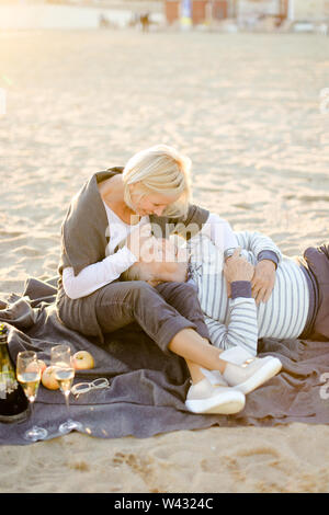 Älterer Mann liegen auf dem Küssen Frau knien und Plaid, Champagner und Früchte auf Sand Strand. Konzept der Rentner auf Picknick und gerne ältere Paare. Stockfoto