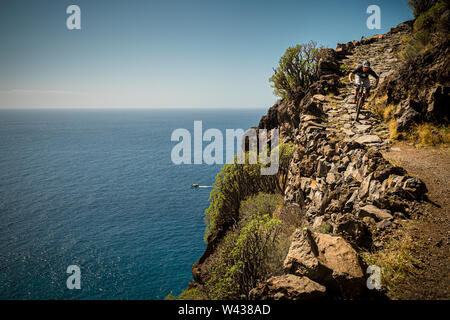 Ein Mountainbiker Fahrten hinunter einen steilen felsigen Pfad auf dem Rand einer Klippe. Ein kleines Fischerboot liegt im Atlantischen Ozean sichtbar unten. Stockfoto