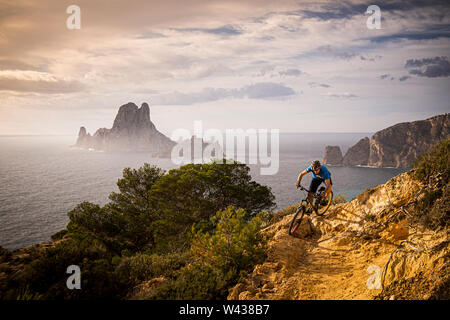 Ein Mann reitet ein Mountainbike entlang einer felsigen Trail an der Westküste von Ibiza. Es Vedrá Insel und das Mittelmeer sind im Hintergrund. Stockfoto
