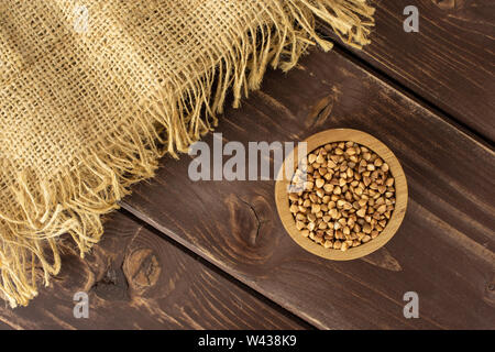 Menge ganze rohe braun Buchweizen Körner in eine hölzerne Schüssel mit einem Stück Jute-stoff flatlay auf braunem Holz Stockfoto