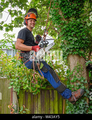 Lächelnde Baum Chirurg oder Baumzüchter mit Kettensäge und Sicherungsseile Arbeit starten Sie einen Baum. Stockfoto