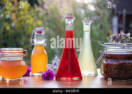 Hausgemachtes Öl aus Johanniskraut Blüten, mit verschiedenen pflanzlichen Sirupe und Blumen im Hintergrund Stockfoto