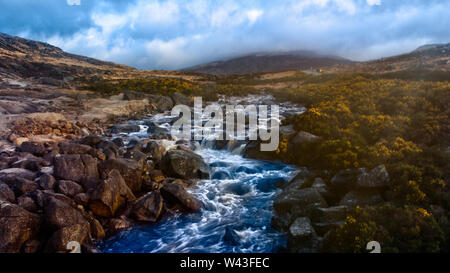 Landschaft Blick auf einen blauen Fluss und Wiese mit gelben Blumen, Berge mit Wolken im Hintergrund umgeben. Sonnenlicht in der linken Seite. Stockfoto