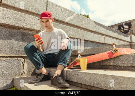 Skateboarder Vorbereitung für das Reiten an der Hauptstraße der Stadt in sonniger Tag. Junger Mann in Sneakers und Kappe mit einem Longboard auf dem Asphalt. Konzept der Freizeitgestaltung, Sport, Extreme, Hobby und Bewegung. Stockfoto