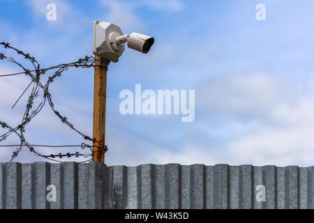 Profilierte blatt Zaun mit billigen Überwachungskamera und Stacheldraht mit selektiven Fokus bei Tageslicht auf blauen Himmel mit Wolken verschwommenen Hintergrund. Stockfoto