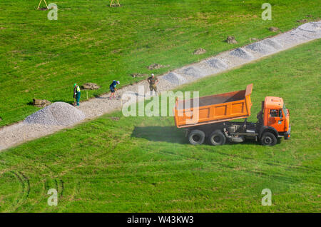 Sankt Petersburg, Russland - Juni 20,2019: Dump Truck KAMAZ gegossen Kies und Wolken aus Staub und fährt los. Die Arbeiter mit Schaufeln. Stockfoto