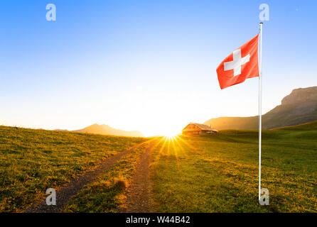 Kleine Straße zu einem Schweizer Chalet oder Bauernhof in der Berglandschaft bei Sonnenaufgang mit Sun Star führenden und eine wehende schweizer Fahne im Vordergrund. Stockfoto