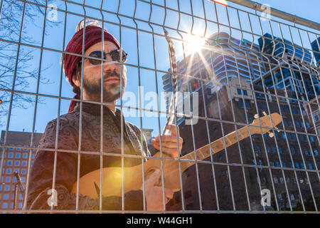 Eine Detailansicht der ein hübscher junger Geistlicher Kerl, hinter einer Kette stehen - einen Zaun in der Nähe von modernen Wolkenkratzern, halten eine kleine Gitarre und symbolische eagl Stockfoto