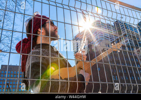 Eine junge Kaukasier Männlich gesehen wird, ein gebürtiger Eagle Feather betrachten und mit einer kleinen Saiteninstrument, hinter einem chainlink fence Downtown. Stockfoto
