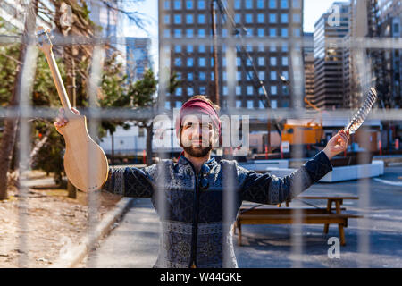 Ein junger kaukasischer Mann gesehen wird hinter einem Maschendrahtzaun in einem städtischen Plaza mit seinen Wolkenkratzern, die im Besitz einer Eagle Feather und akustischen Instruments. Stockfoto