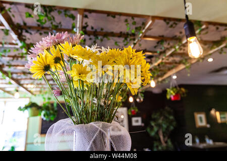 Eine Detailansicht der gelben und frische Blumen in einem modernen Vase auf eine Tabelle innerhalb einer trendigen eco-freundliches Restaurant. Ivy hängen von Holzbalken im Hintergrund. Stockfoto