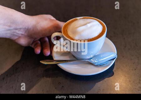 Eine Detailansicht an Hand einer Person erreichen für einen frisch zubereiteten Latte in einem Coffeeshop. Aromatische Frühstück trinken im Hotel serviert. Stockfoto