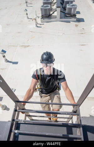 Hvac Tech das Tragen von Schutzausrüstung, hinunter klettern eine Leiter auf einem Dach mit Kondensatoren im Hintergrund. Stockfoto