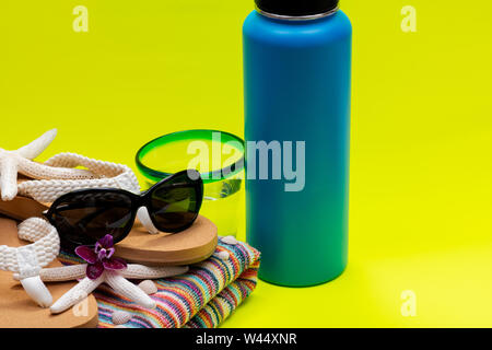 Blau isolierte Edelstahl Flasche, Strandtuch, Flip Flops, blaue Felge Glas mit Wasser und Schwarze Sonnenbrille auf gelb wie Sommer Urlaub Thema. Stockfoto