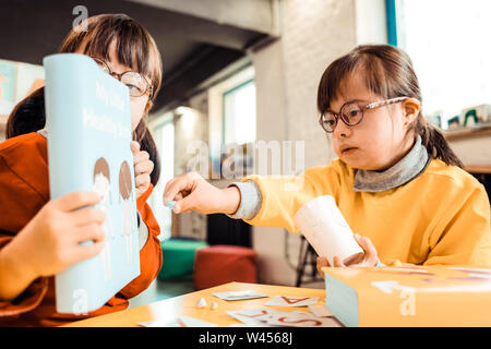 Neugierig kid mit gebunden Haar zeigt auf einem hellen Abdeckung des Lernens Buch Stockfoto
