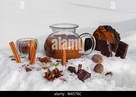 Zimtstangen, Kräuter und Gewürze in der Nähe gesehen, stehend im Schnee mit einem Glas Urne mit chaga Chai Tee gefüllt, einem alten Rezept in der alternativen Medizin verwendet Stockfoto