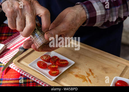 Eine Nahaufnahme auf die Hände eines älteren Mannes, mit einer manuellen Schleifer zu Saison in Scheiben geschnittene Tomaten mit getrockneten Kraut Flocken während ein natürliches Lebensmittel Messe. Stockfoto