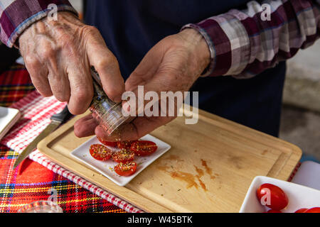 Aus der Nähe zu sehen. eine Person mit Händen zu streuen getrocknete Kraut Flocken über geschnittene Pflaumen Tomaten während ein homegrown Essen Messe, die Vorbereitung von Proben für die Verbraucher. Stockfoto