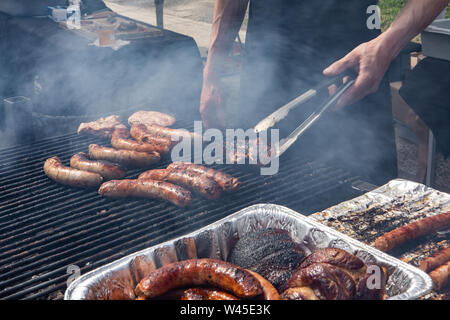 Eine Nahaufnahme Blick auf ein warmes Essen Anbieter kochen Würstchen und Koteletts über einem heißen Grill mit aufsteigenden Dampf, Küchenchef verwendet Zangen Fleisch auf lokaler landwirtschaftliche Messe auf Flip Stockfoto