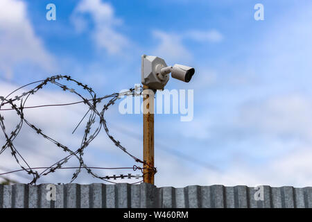 Profilierte blatt Zaun mit billigen Überwachungskamera und Stacheldraht mit selektiven Fokus bei Tageslicht auf blauen Himmel mit Wolken verschwommenen Hintergrund. Stockfoto