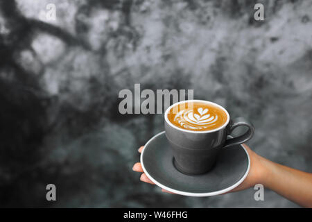 Die Frau in der Hand grau Tasse frischen Cappuccino Holding auf dem Hintergrund von grauem Beton Zement texturierte Wand. Trendy hipster Cafe Design. Stockfoto