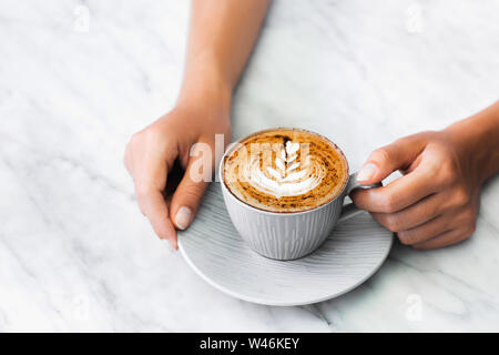 Tasse frischen Kaffee Cappuccino in der Frau die Hände auf weißem Marmor tisch trendy Hintergrund. Klassische latte Kunst und Schokolade auf Schaum. Leeren Platz für Text, Stockfoto