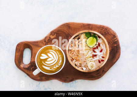 Frischen leckeren Smoothie Schüssel mit Bananen, Müsli, Chia Samen, Goji Beeren, Kokosraspeln und Schneiden von Kalk. Tasse Cappuccino. Gesunde vegetaria Stockfoto