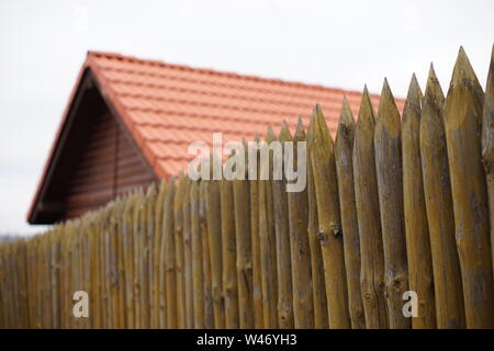 Ein Zaun aus spitzen Holzpfählen vor dem Hintergrund der ein Holzhaus mit einem roten Ziegeldach und einen grauen Himmel. Stockfoto