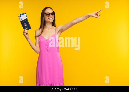 Schlanke Mädchen in ein rosa Kleid und Sonnenbrille holding Reisepass, Flugticket und weg zeigend auf gelbem Hintergrund Stockfoto