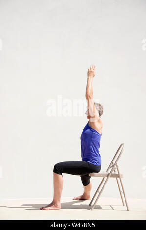 Reifer senior Frau practing Sanfte restorative Yoga in einem weißen Hintergrund darstellt. Sie hat mit erhobenen Händen, während er auf eine spezielle Therapie Stuhl. Stockfoto