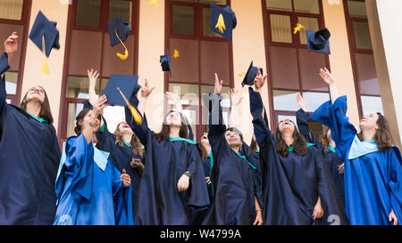 Hochschulabsolventen werfen Graduierung Hüte in der Luft. Gruppe der glücklichen Absolventen der akademischen Kleider in der Nähe der Universität. Junge Studenten Kleid Stockfoto