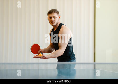 Mann mit Ping pong Schläger vorbereiten, einen Ball zu schlagen, Training im Innenbereich. Männliche Person in Sportbekleidung, Training im Tischtennis club Stockfoto