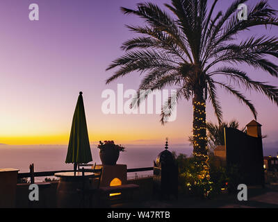 Lila goldene Abendstimmung an der Nordküste von Teneriffa, mit einer großen Palme umgeben von leichten Kette auf einer Aussichtsterrasse mit Blick auf den Stockfoto