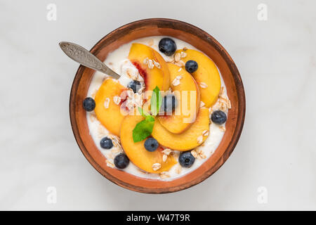 Joghurt mit Pfirsichen, Hafer, Blaubeeren, Honig und Minze in eine Schüssel geben und mit einem Löffel für gesundes Frühstück auf weißem Hintergrund. top View Stockfoto