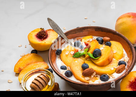 Joghurt mit Pfirsichen, Hafer, Blaubeeren, Honig und Minze in eine Schüssel geben und mit einem Löffel für gesundes Frühstück auf weißem Hintergrund. Nach oben Schließen Stockfoto