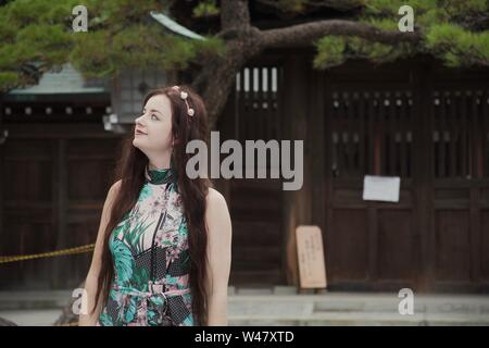 Eine brunette, Kaukasier, langhaarigen, modischen hippie Mädchen mit Blumen (Flower crown) steht lächelnd vor einer Kiefer in einem japanischen Cour Stockfoto