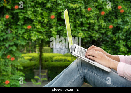 Junge schöne Frau im Freien in einem öffentlichen Park. Arbeiten am Laptop im Freien. Zugeschnittenes Bild der weiblichen Arbeiten am Laptop während der Sitzung in einem Park. Stockfoto