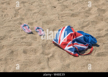 Union Jack Strandtasche & Flip-Flops am Sandstrand - für 2021 Aufenthalte in Großbritannien, Urlaub zu Hause, Aufenthalt in Cornwall, Badeurlaub, Flip-Flop-Schuhe. Stockfoto