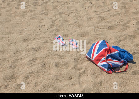 Union Jack Strandtasche & Flip-Flops am Sandstrand - für 2021 Aufenthalte in Großbritannien, Urlaub zu Hause, Aufenthalt in Cornwall, Badeurlaub, Flip-Flop-Schuhe. Stockfoto