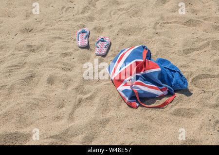 Union Jack Strandtasche & Flip-Flops am Sandstrand - für 2021 Aufenthalte in Großbritannien, Urlaub zu Hause, Aufenthalt in Cornwall, Badeurlaub, Flip-Flop-Schuhe. Stockfoto