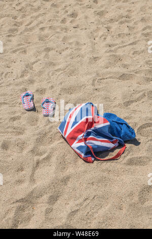 Union Jack Strandtasche & Flip-Flops am Sandstrand - für 2021 Aufenthalte in Großbritannien, Urlaub zu Hause, Aufenthalt in Cornwall, Badeurlaub, Flip-Flop-Schuhe. Stockfoto